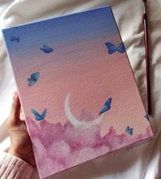 a person holding up a painting with blue butterflies flying over the moon and pink clouds