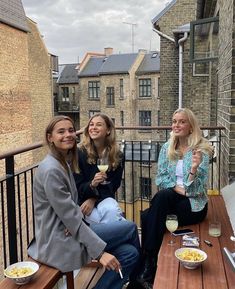 three women sitting at a table with food and drinks on the outside balcony overlooking brick buildings