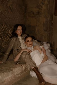 two women sitting on the floor in front of a stone wall, one wearing a white dress