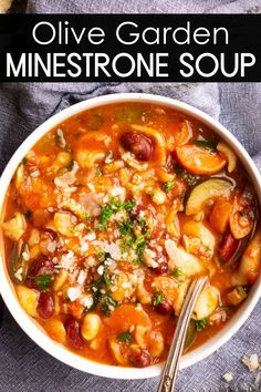 A bowl of minestrone soup filled with a mix of vegetables, beans, pasta, and herbs, topped with grated cheese. A spoon is resting inside the bowl, and its placed on a dark fabric surface.