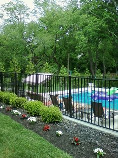 a fenced in swimming pool with chairs and umbrellas next to it, surrounded by lush green trees