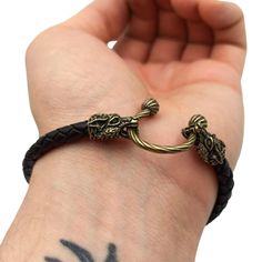 a person with a tattoo on their arm holding onto a black leather bracelet that has two gold dragon charms attached to it