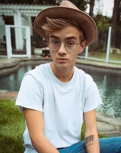 a young man wearing glasses and a hat sitting on the grass next to a pool