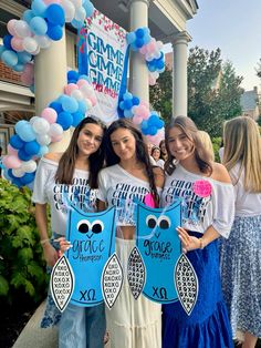 three girls holding up signs and balloons in front of a building that says gimme omme