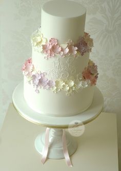 a three tiered white cake with pink and purple flowers on the top, sitting on a pedestal