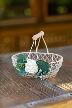 a wire basket with white flowers and green leaves on it sitting on top of a wooden table