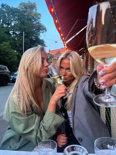 two women sitting at an outdoor table drinking wine and talking to each other while holding glasses in their hands