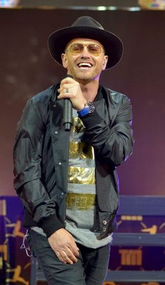 a man in a black hat and jacket holding a microphone with his right hand to his chest