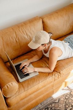 a woman laying on a couch using a laptop computer while wearing a baseball cap and jeans