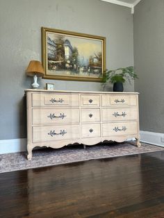 a white dresser sitting on top of a hard wood floor next to a painting and lamp