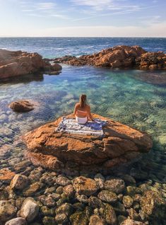 a woman sitting on top of a rock next to the ocean with a towel in her lap