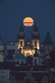 the full moon is setting over a city with tall buildings and spires in front of it