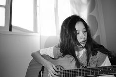 a woman sitting in front of a window while playing an acoustic guitar with long hair