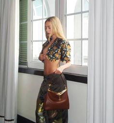 a woman standing in front of a window holding a brown purse