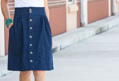 a woman standing on the sidewalk wearing a blue skirt and white tank top with buttons