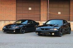 two black sports cars parked next to each other in front of a brick building with garage doors