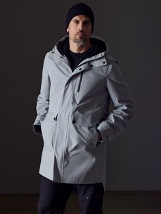 Front body view of man wearing Anders Rain Jacket in Sky Grey from AETHER Apparel. Weatherproof Long Coat For Workwear, Modern Waterproof Raincoat For Workwear, Waterproof Long Coat For Work, Waterproof Long Coat For Workwear, Weatherproof Long Coat For Rainy Weather, Modern Raincoat With Detachable Hood And Long Sleeves, Techwear Outerwear With Detachable Hood For Work, Modern Nylon Outerwear, Modern Nylon Outerwear For Rainy Weather