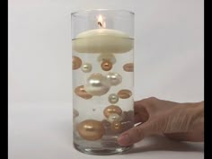 a hand is holding a candle in a glass filled with pearls and other things that are floating around