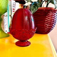 a red glass vase sitting on top of a yellow table next to a green plant