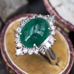 This magnificent ring is centered with one (1) oval cabochon cut natural emerald weighing 5.18 carats and set into a four-prong setting. The emerald is bordered with eight (8), prong set, square step cut diamonds and twelve (12), prong set, round brilliant cut diamonds. The ring measures 23.1mm at the top, rises 12.2mm above the finger, tapering to 3.8mm wide and 1.9mm thick at the base of the shank. This ring is currently a size 7.75. Luxury Oval Emerald Birthstone Ring, Fine Jewelry Emerald Oval Cabochon Ring, Fine Jewelry Emerald Ring With Oval Cabochon, Luxury Emerald Ring With Oval Cabochon And 17 Jewels, Luxury Oval Cabochon Emerald Ring With 17 Jewels, Emerald Oval Cabochon Ring In Fine Jewelry Style, Classic Cabochon Emerald Ring, Elegant Emerald Ring With Cabochon Cut, Elegant Cabochon Emerald Ring