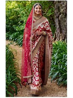 a woman in a red and gold sari standing on a dirt path with trees behind her