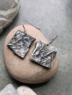 VORA GRISOS Earrings Reticulated metal Hand chiselled Patinated silver Solid silver ear hooks Delivered in its red and gold pouch Maintains like classic silver with a dry and soft cloth, or with silverware if necessary. I used bronze plates that I had in my workshop and I sawed small square shapes. I then heated them with a torch to a certain temperature, so that the metal melted but not completely and I managed my flame to give them this textured appearance. This is called cross-linking. I then reworked them, using small goldsmith chisels of abstract shapes. The metal was then completely stripped using chemical processes. The silvering was made by electrolysis in my workshop in Nice. Their shape is unique, and the two loops are different in appearance which gives them a more aesthetic. Yo Earrings Geometric, Jewellery Earrings, Earrings Metal, Bronze Metal, Metal Hand, Recycled Metal, Ear Hook, Metal Earrings, Chic Woman