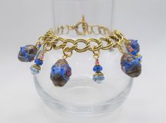 "This handmade gold tone charm bracelet by Lauriechacha features vintage Venetian Murano wedding cake motif and aventurine lamp work glass beads, Swarovski beads, vintage glass beads, TierraCast antique gold heishi beads and lily beadcaps, wire wrapped on a vintage double oval curb chain. The colors are Light Sapphire, Sapphire, Rose and Light Rose. It has an Heirloom antique gold TierraCast toggle clasp. The bracelet measures approximately 8-1/4\" in length by 1-1/2\" in width and weighs 41 gra Gold Beaded Wedding Charm Bracelet, Handmade Gold Charm Bracelet For Wedding, Gold Glass Jewelry For Celebration, Celebration Gold Glass Jewelry, Gold Beaded Charm Bracelet For Wedding, Gold Beaded Bracelets With Dangling Beads, Gold Bracelets With Dangling Beads For Gift, Gold Wedding Charm Bracelet With Round Beads, Gold Glass Jewelry With Dangling Beads