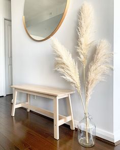 a wooden bench sitting next to a tall white plant in a vase on top of a hard wood floor