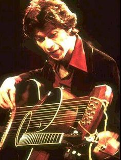 a man playing an acoustic guitar while wearing a black jacket and red shirt with his hands on the strings