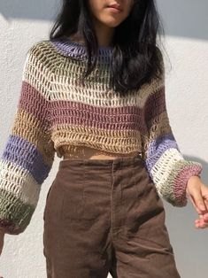 a woman wearing brown pants and a multicolored crochet sweater is standing in front of a white wall