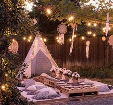 an outdoor dining area with lights strung over the table and pillows on the ground, in front of a wooden fence