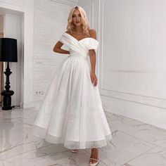 a woman wearing a white dress standing in front of a wall and posing for the camera