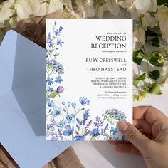 a person holding up a wedding card with blue flowers on it and white envelope in the background