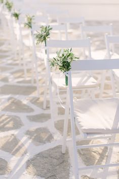 rows of white folding chairs with greenery tied to them on the beach for an outdoor wedding ceremony