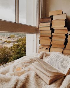 a pile of books sitting on top of a bed next to a window with a candle