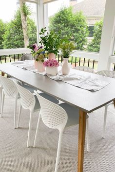 a dining table with white chairs and flowers in vases on the top, next to an enclosed porch