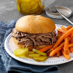 a roast beef sandwich with carrots and pickles on a plate next to a jar of pickles