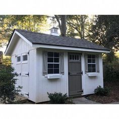 a small white shed with windows and shutters on the side, in front of some trees