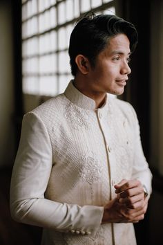 a man standing in front of a window wearing a white suit and holding his hands together