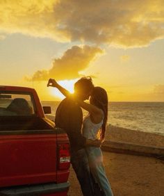 a man and woman standing next to a red truck on the beach at sunset with their arms around each other