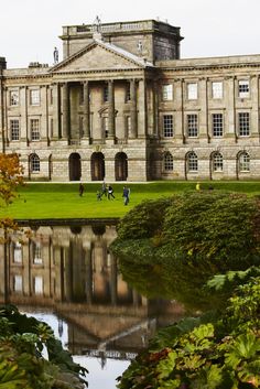 two people are walking in front of an old building with water and greenery around it