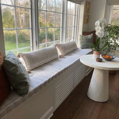 a window seat with pillows on it in front of a table and vase filled with flowers