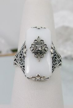 White Glass & Inset Gemstone (CZ, Moissanite, or Diamond) Intricate Filigree Ring Grace Design#233 Custom Made I now offer this lovely Antique Art Deco/Edwardian design ring in sterling silver. This gorgeous ring is set with a stunning floral filigree embellished white glass/resin with a centered inset 1mm white round cut CZ, Lab Moissanite, or Natural Diamond (Choose option above). The glass is 18mm Long by 19mm Wide. The ring sits 9mm North South on the finger and 10mm East West on the finger. Fine Jewelry White Sterling Silver Rings, White Sterling Silver Fine Jewelry Rings, Antique White Ring With Center Stone, White Sterling Silver Ring Jewelry, Classic White Jewelry With Diamond Accents, Ornate Formal Jewelry With Stone Setting, Ornate Jewelry With Stone Setting For Formal Occasions, Elegant White Filigree Ring With Intricate Design, Ornate Jewelry For Formal Occasions With Stone Setting