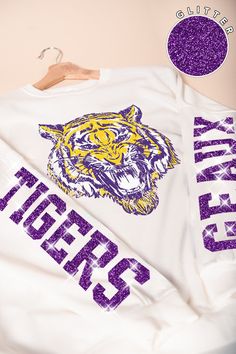 Our Glitter Geaux Tigers sweatshirt is perfect for a gameday in Tiger Stadium! This is an oversized sweatshirt that comes in sizes S/M (2-6) and M/L (8-12). This trendy long-sleeve shirt features a bold tiger graphic in vibrant yellow and purple on the front, making it a standout piece for any casual wardrobe. The shirt also showcases "Tigers" in glittery purple lettering along the sleeve, adding a touch of sparkle and school spirit. Perfect for game days, casual outings, or lounging in style, t Game Day Graphic Print Long Sleeve Sweatshirt, Oversized Sweatshirt With Ribbed Cuffs For Game Day, Fall Fan Gear Graphic Sweatshirt, Fall Fan Gear Sweatshirt With Graphic Print, Fan Gear Graphic Print Sweatshirt For Fall, Oversized Sweatshirt For Game Day And Sports Season, Oversized Sweatshirt For Game Day, Oversized Varsity Sweatshirt For Game Day, Varsity Long Sleeve Sweatshirt For Game Day