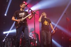 two men standing on stage with microphones in front of them and one holding a guitar