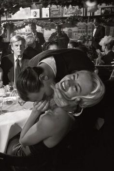 black and white photograph of woman kissing man's cheek at table with other people in background