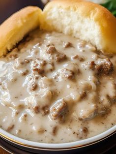 a bowl filled with meat and gravy on top of a table next to a roll