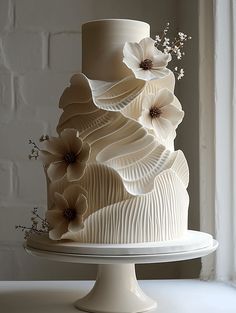 a three tiered cake with white icing and flowers on top, sitting in front of a window