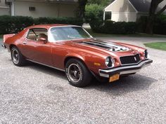 an orange muscle car parked in a driveway next to some bushes and trees with a house in the background