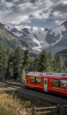 a red train traveling through a lush green forest covered mountain side under a cloudy sky